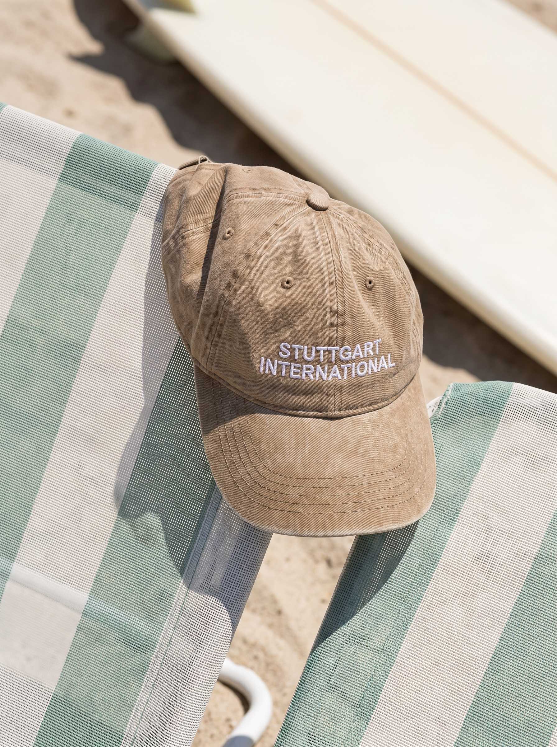 Verwaschen hellbraune „Stuttgart International“-Cap liegt auf einem gestreiften Liegestuhl in der Sonne.