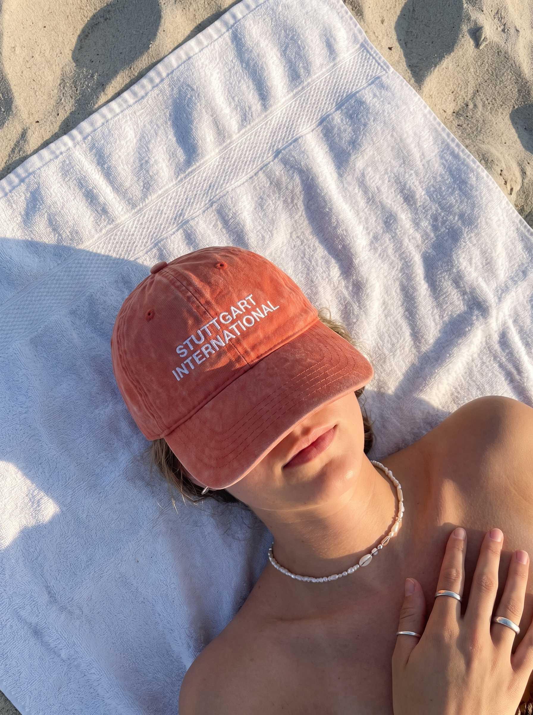Verwaschen rote „Stuttgart International“-Cap bedeckt das Gesicht einer Person, die auf einem Handtuch am Strand liegt.