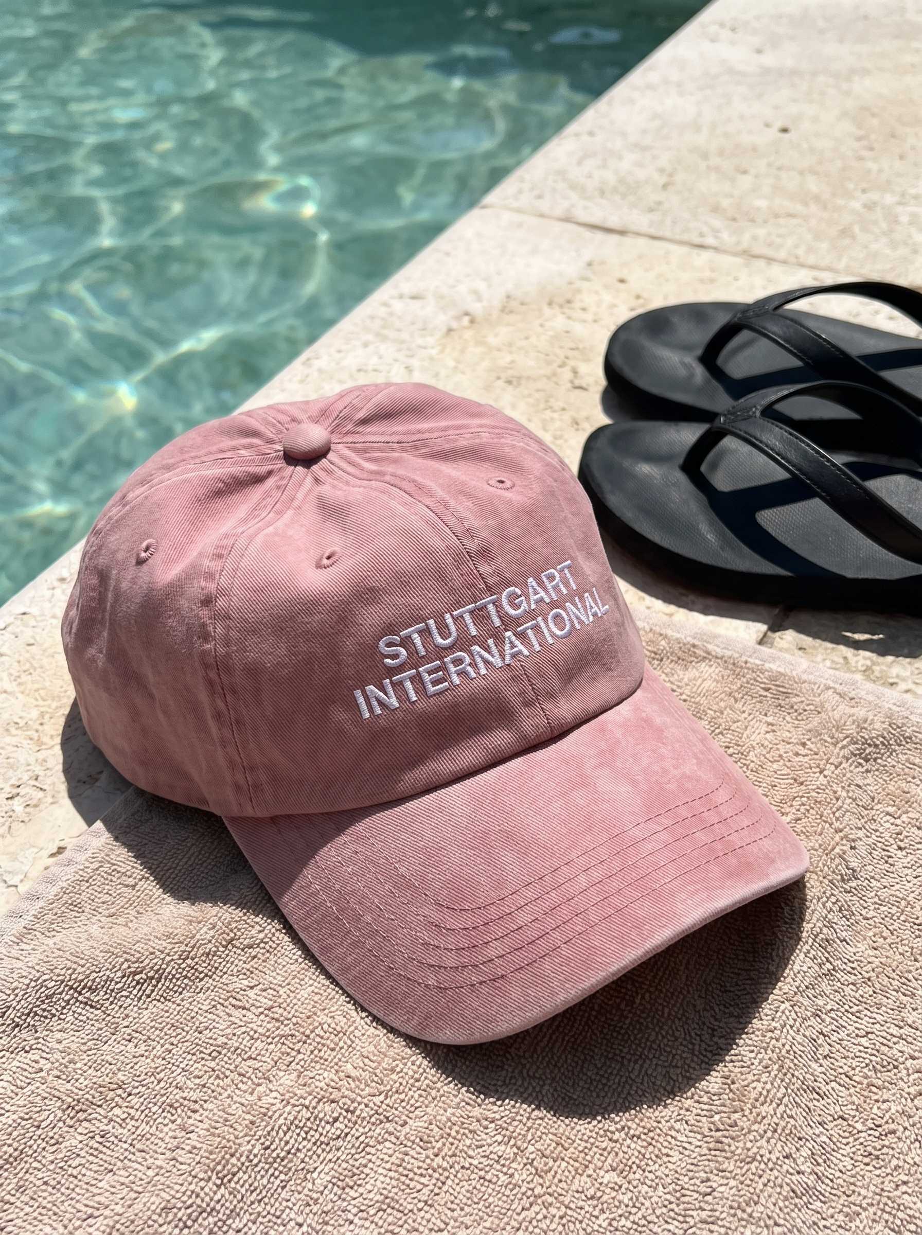Verwaschen rosafarbene „Stuttgart International“-Cap liegt neben schwarzen Sandalen auf einem Handtuch am Poolrand.