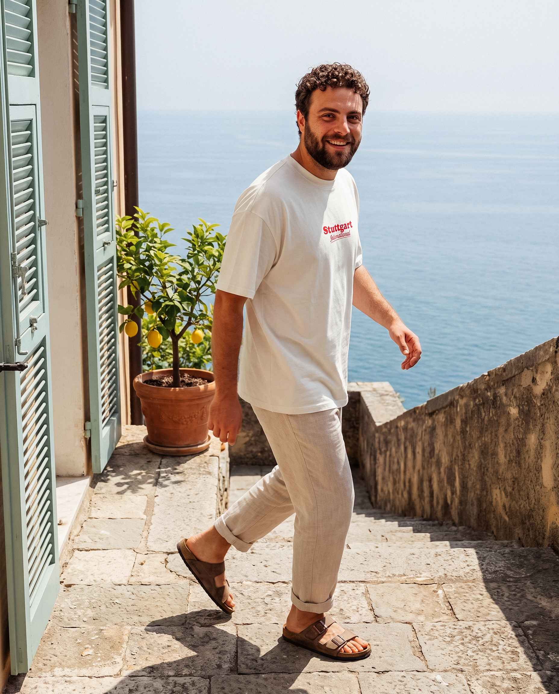 Mann mit Stuttgart Shirt "International" steht seitlich zur Kamera und schaut in die Kamera. Hinter ihm das Meer.