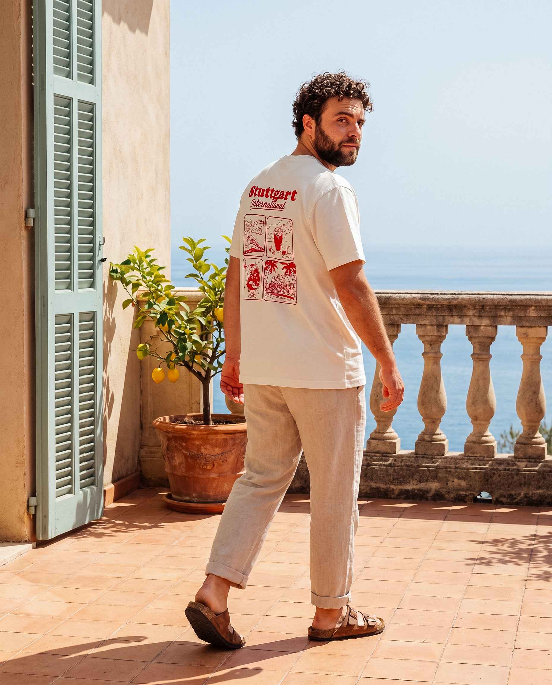 Mann mit Stuttgart Shirt "International" steht mit dem Rücken zur Kamera und schaut in die Kamera. Er steht auf einer Terasse vor einer Steinbalustrade.