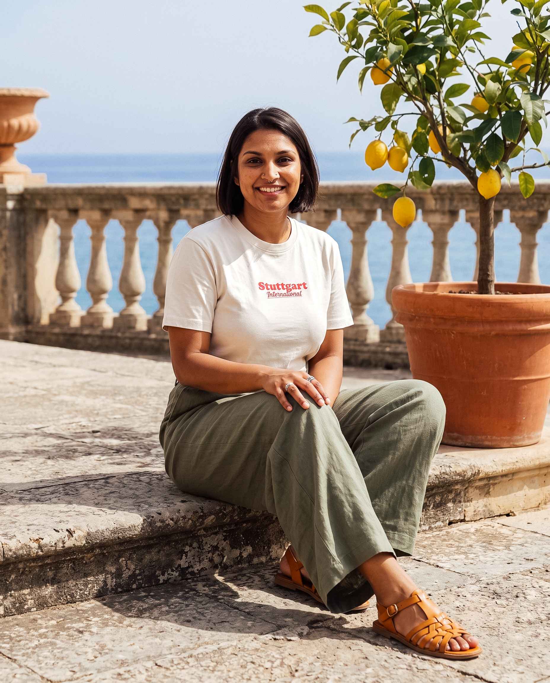 Frau sitzt auf dem Boden neben einem Zitronenbaum und trägt dabei das Stuttgart Shirt "International" und schaut dabei fröhlich in die Kamera.